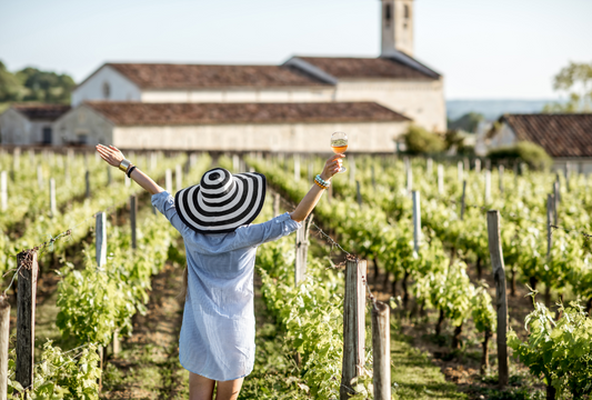 A woman wearing a hat in a vineyard celebrating and enjoying a glass of wine.